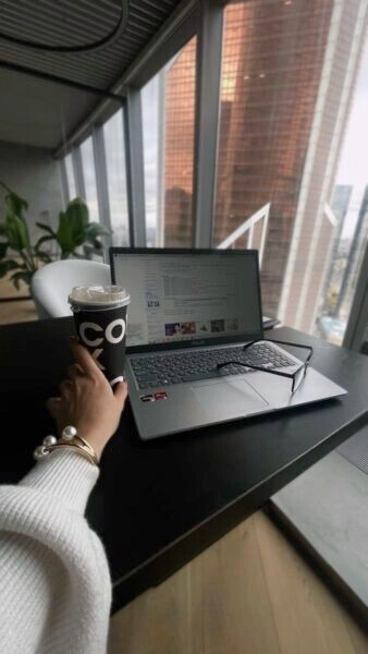 An image is clicked from the person's angle, showing her desk where there's a laptop on which stands a pair of glasses, and near the laptop, she shows a cafe-style coffee cup that she's holding, altogether giving a sense that she's working in an office.