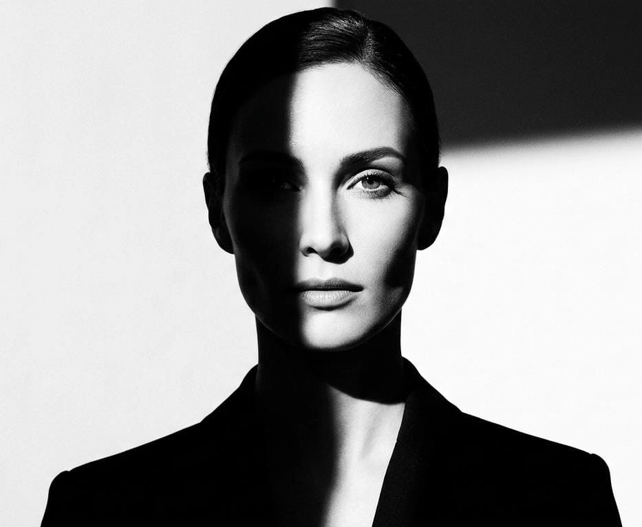 A monochrome image of a lady looking straight towards the camera. She wears a formal suit. Seems serious and stands with confidence and elegance. The background is filled with a plain white wall and a shadow cast by the female.
